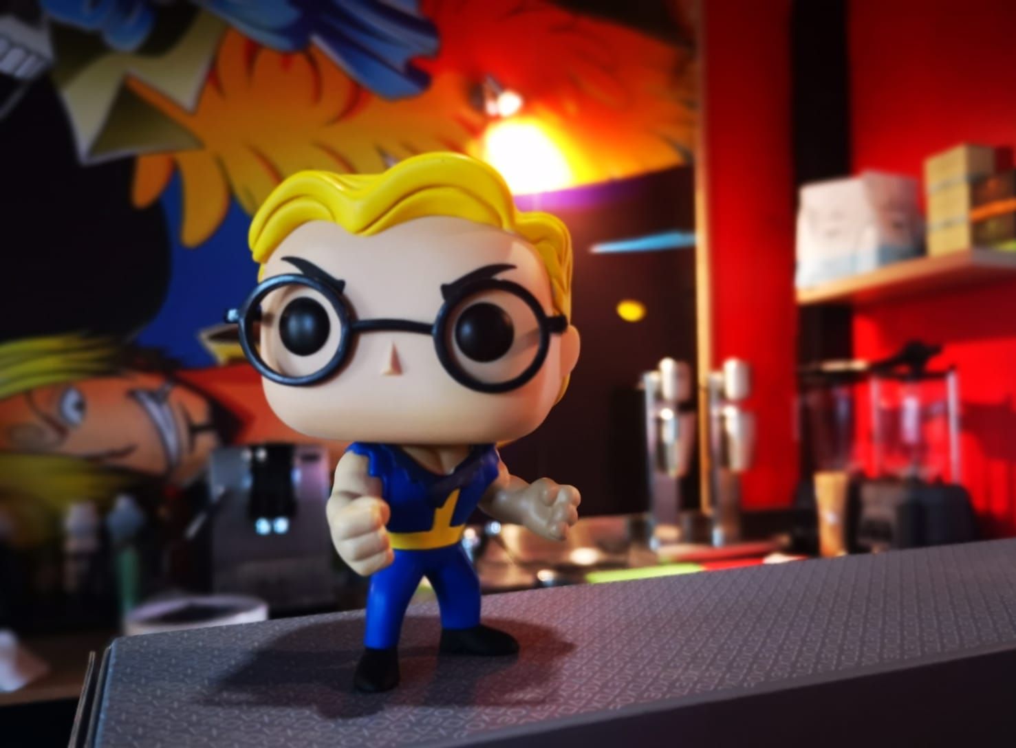 Photo illustrative d’un funko pop représentant un habitant des bunker dans Fallout avec le bar du café en arrière plan