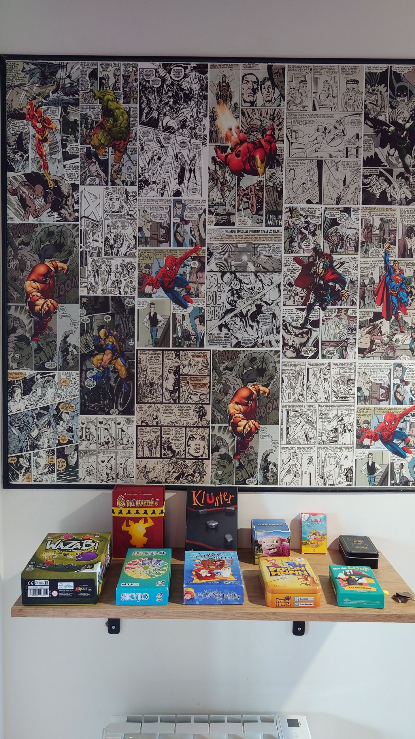 Photo d’un cadre au mur représentant un patchwork de page de comics. Il y a des jeux de société sur une étagère en dessous.
